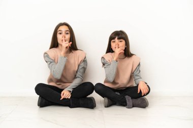 Beyaz arka planda izole bir şekilde yerde oturan küçük kız kardeşler sessiz bir jest işareti gösteriyorlar. Parmağını ağzına sokuyorlar.