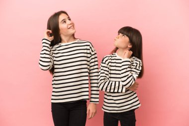 Küçük kız kardeşler pembe arka planda izole edilmiş kafalarını kaşırken bir fikir düşünüyorlar.