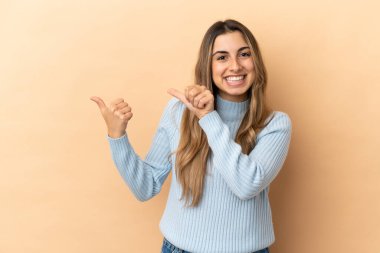 Beyazlı genç bir kadın bej arka planda izole edilmiş bir şekilde bir ürün sunmak için kenarı işaret ediyor.