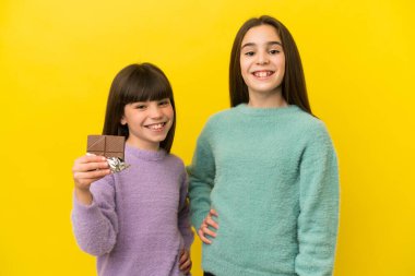 Sarı arka planda izole edilmiş küçük kız kardeşler çikolata tableti alıyor ve mutlular.