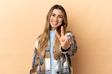 Bej arka planda izole edilmiş genç beyaz kadın gülümsüyor ve zafer işareti gösteriyor.