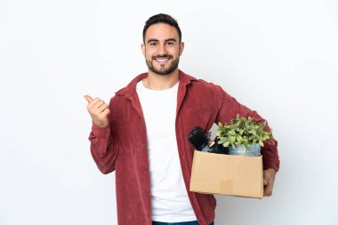 Beyaz arka planda izole edilmiş eşyalarla dolu bir kutuyu alıp bir ürün sunmak için kenarı işaret eden genç beyaz bir adam hamle yapıyor.