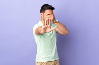 Mor arka planda izole edilmiş genç yakışıklı adam dur hareketi yapıyor ve yüzünü kapatıyor.