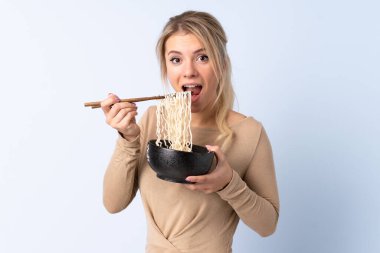 Sarışın kadın, izole edilmiş mavi arka planda elinde çubuklarla bir kase erişte tutuyor ve onu yiyor.