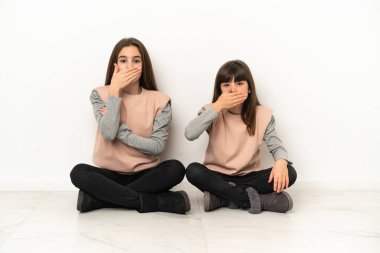 Beyaz arka planda izole bir şekilde yerde oturan küçük kız kardeşler uygunsuz bir şey söylediği için ağızlarını ağızla kapatıyorlar.