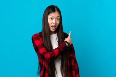 Genç Çinli kız izole edilmiş mavi arka planda şaşırmış ve işaret ediyor.