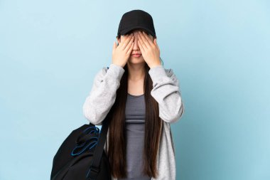 İzole edilmiş mavi arka planda spor çantalı genç Çinli kadın elleriyle gözlerini kapatıyor.