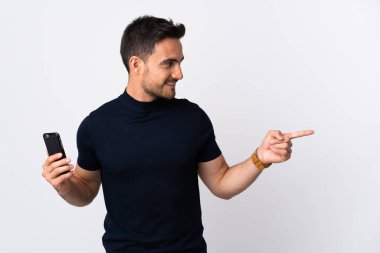 Beyaz arka planda izole edilmiş cep telefonu kullanan genç beyaz bir adam bir ürün sunmak için kenarı işaret ediyor