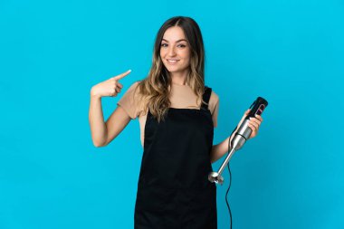 Mavi arka planda izole edilmiş el blenderi kullanan Romen kadın başparmak hareketi yapıyor