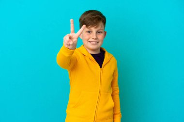 Mavi arka planda izole edilmiş küçük kızıl çocuk gülümsüyor ve zafer işareti gösteriyor.