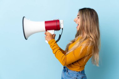 İzole edilmiş mavi arka planda genç İspanyol kadın megafondan bir şey duyurmak için bağırıyor.