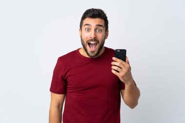 Sakallı genç yakışıklı adam beyaz arka planda izole edilmiş, şaşırmış ve şok olmuş bir yüz ifadesiyle cep telefonu kullanıyor.