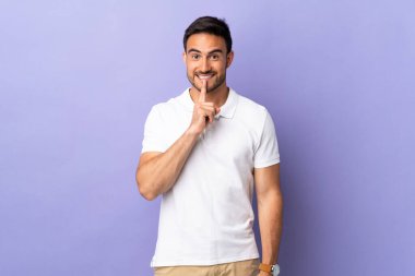 Mor arka planda izole edilmiş genç yakışıklı bir adam sessizlik işareti gösteriyor. Parmağını ağzına sok.