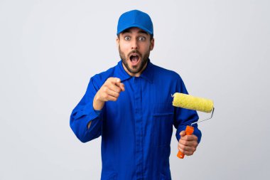 Beyaz arka planda izole edilmiş bir boya silindiri tutan ressam şaşırmış ve ön tarafı işaret ediyor.