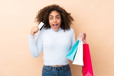 Genç Afro-Amerikalı kadın bej arka planda izole edilmiş elinde alışveriş torbalarıyla şaşırmış.