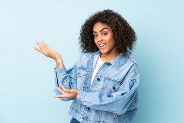 Genç Afrikalı Amerikalı kadın mavi arka planda izole edilmiş. Davet etmek için ellerini yana uzatıyor.