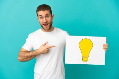 Sarışın, soyutlanmış, mavi arka planda, elinde ampul simgesi olan bir levha tutan yakışıklı bir adam.