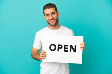 Yakışıklı sarışın, izole edilmiş mavi arka plan. Elinde mutlu bir ifadeyle açık yazılı bir pankart var.