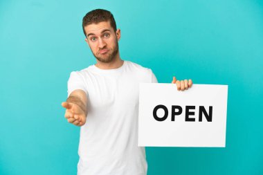 Yakışıklı sarışın, izole edilmiş mavi arka planda elinde bir pankartla bir anlaşma yapıyor.