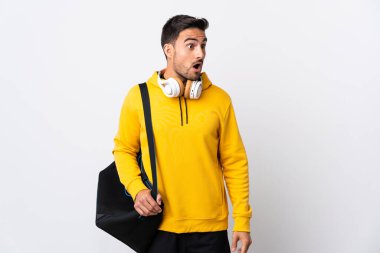 Beyaz arka planda izole edilmiş spor çantalı genç adam yan tarafa bakarken sürpriz jest yapıyor.