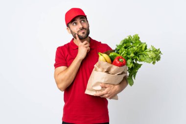 Sakallı genç bir adam elinde bir çanta dolusu sebzeyle beyaz arka planda izole edilmiş bir fikir düşünüyor.