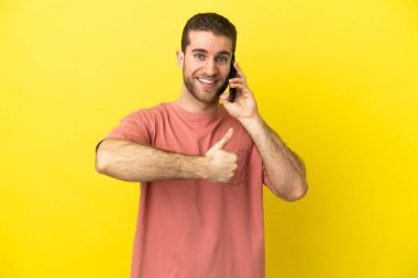 Sarışın yakışıklı adam, izole edilmiş arka planda cep telefonu kullanıyor. Başparmak işareti yapıyor.