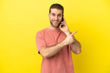 Yakışıklı sarışın adam izole edilmiş arka planda cep telefonu kullanıyor. Ürün sunmak için kenarı işaret ediyor.
