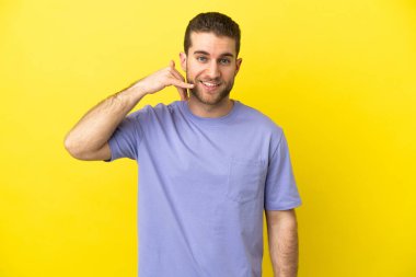 Sarışın yakışıklı adam izole edilmiş sarı arka planda telefon hareketlerinde bulunuyor. Bana geri dönüş işareti ver.