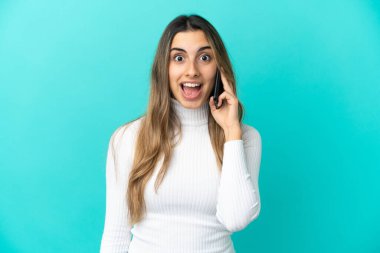 Mavi arka planda izole edilmiş cep telefonu kullanan beyaz bir kadın. Sürpriz bir yüz ifadesi var.