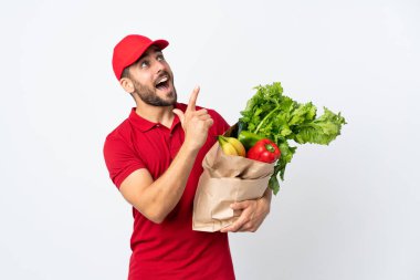 Sakallı genç adam elinde sebzelerle dolu bir çanta tutuyor beyaz arka planda, işaret parmağıyla işaret ediyor. Harika bir fikir.
