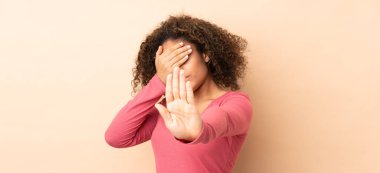 Genç Afro-Amerikalı kadın bej arka planda izole edilmiş. Durup jest yapıyor ve yüzünü kapatıyor.