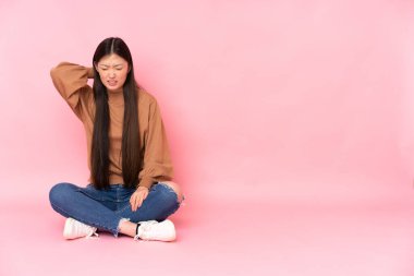 Genç Asyalı kadın pembe arka planda boynu ağrıyan bir şekilde yerde oturuyor.