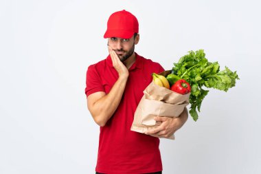 Sakallı genç adam elinde sebze dolu bir çanta tutuyor. Beyaz arka planda izole edilmiş mutsuz ve hayal kırıklığına uğramış.