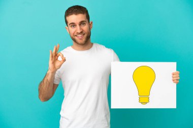 İzole edilmiş mavi arkaplanı olan yakışıklı sarışın adam üzerinde ok işareti olan ampul simgesi olan bir levha tutuyor.