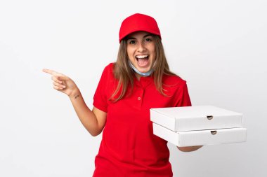 Pizza teslimatı yapan kadın elinde pizza tutuyordu. Koronavirüsten korunuyordu. İzole edilmiş beyaz duvarın üzerinde maskesi vardı.