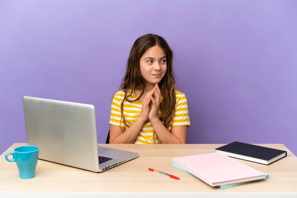 Little student girl in a workplace with a laptop isolated on purple background scheming something