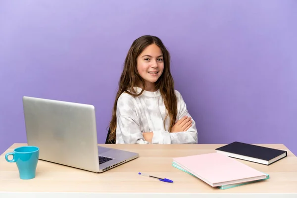 Little student girl in a workplace with a laptop isolated on purple background with arms crossed and looking forward