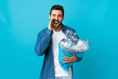 Mavi arka planda izole edilmiş, ağzı açık bağırarak geri dönüşüm için plastik şişelerle dolu bir çanta tutan genç beyaz bir adam.