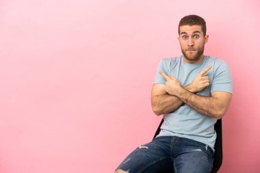 İzole edilmiş pembe arka planda bir sandalyede oturan genç adam yan taraftaki kuşkuları işaret ediyor.