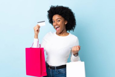 Genç Afrikalı Amerikalı bir kadın mavi arka planda izole edilmiş. Elinde alışveriş çantaları ve bir kredi kartı var.
