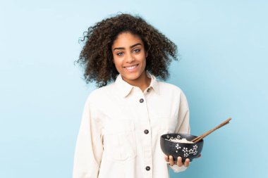 Mavi arka planda izole edilmiş genç Afrikalı Amerikalı kadın elinde çubuklarla elinde bir kase erişte tutarken gülümsüyor.