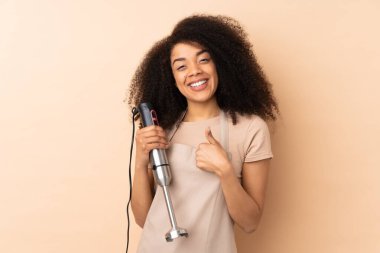 Genç Afrikalı Amerikalı kadın bej arka planda izole edilmiş el blenderi kullanıyor.