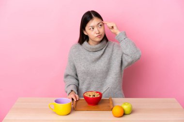 Genç Çinli bir kız kahvaltı yapıyor. Çılgınlık jesti yapıyor. Parmağını kafanın üstüne koyuyor.