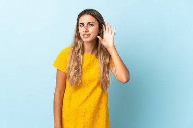 İzole edilmiş mavi arka planda genç bir İspanyol kadın. Kulağına elini sürerek bir şeyler dinliyor.