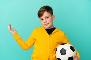 Kızıl saçlı çocuk mavi arka planda izole bir şekilde futbol oynuyor. Davet etmek için ellerini yana açıyor.
