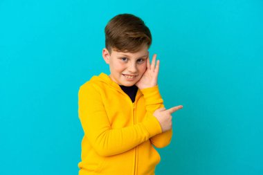 Mavi arka planda izole edilmiş küçük kızıl saçlı çocuk bir ürün sunmak ve bir şeyler fısıldamak için kenarı işaret ediyor.