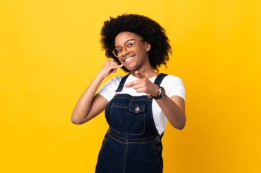 Sarı arka planda izole edilmiş genç Afrikalı Amerikalı bir kadın telefon hareketi yapıyor ve ön tarafı işaret ediyor.