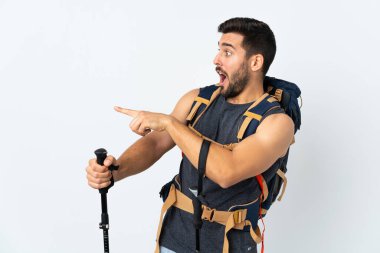Genç dağcı, büyük bir sırt çantası ve beyaz arka planda izole edilmiş yürüyüş direkleri ile parmağı yan tarafı işaret ediyor.