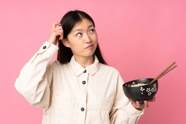 Pembe arka planda izole edilmiş genç Asyalı bir kadın kafasında yemek çubuklarıyla bir kase erişte tutarken...