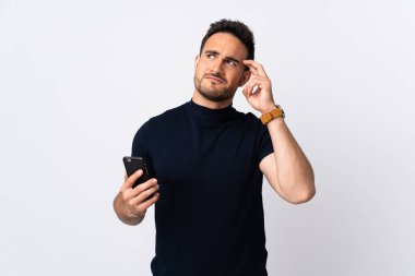 Beyaz arka planda izole edilmiş cep telefonu kullanan Kafkasyalı genç adamın şüpheleri ve kafası karışmış yüz ifadesi var.